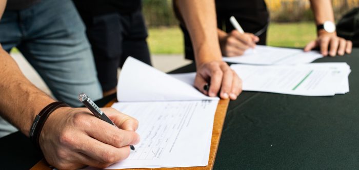 Close up of people's hands signing a form on a wooden clipboard